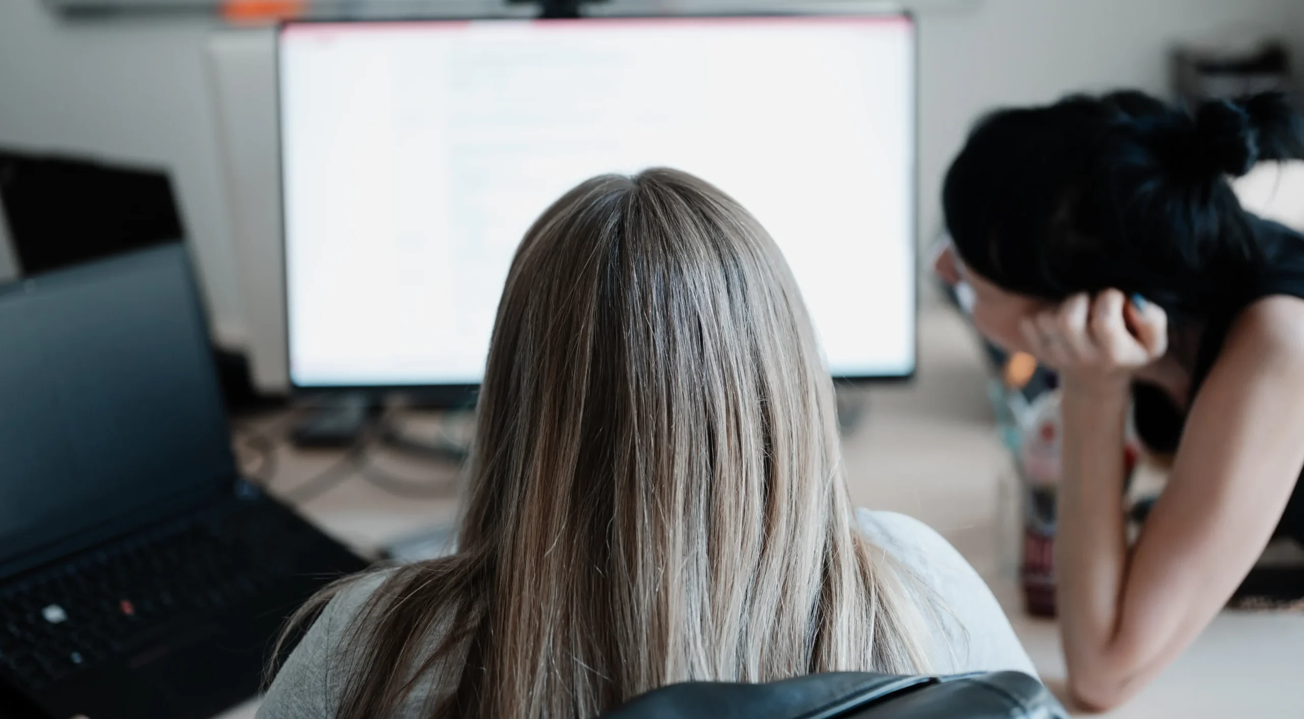 Zwei Mitarbeiterinnen sitzen mit dem Rücken zur Kamera gemeinsam vor einem Bildschirm